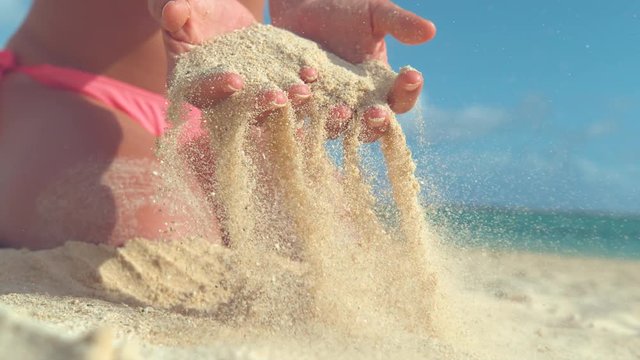 SLOW MOTION, CLOSE UP: Unrecognizable young woman in pink bikini scatter white sand across the sunny shore. Playful girl lifts up a handful of sand and lets it get swept away in the cool ocean breeze.