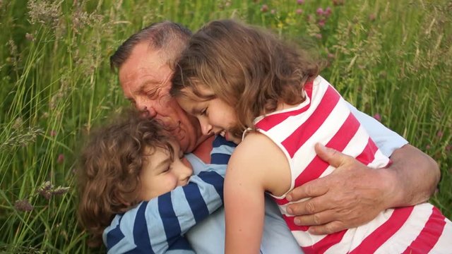 Grandfather and grandchildren, a boy and a girl, a brother and a sister rest on a summer glade. Grandchildren gaily hug grandfather, lay in the grass and laugh. Happy Family Vacation