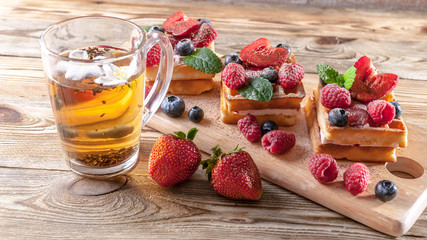 Cup of tea and belgian waffles with strawberry jam, fresh strawberries, raspberries, blueberries and chocolate chips on a wooden table. Homemade baking. Tasty breakfast