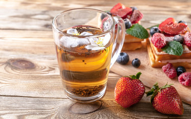 Cup of tea and belgian waffles with strawberry jam, fresh strawberries, raspberries, blueberries and chocolate chips on a wooden table. Homemade baking. Tasty breakfast