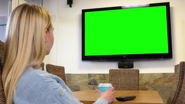 A woman sits at a table in a living room and watches a TV with a green screen