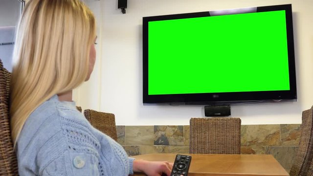 A woman sits at a table in a living room and switches channels on a TV with a green screen