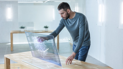 In the Near Future Stylish Man Using His High-Tech Computer with Transparent Display and Checks Useful Work Related Statistics. He Works in the Ultramodern Light Office.