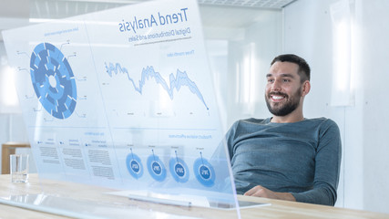 In the Near Future Successful Smiling Trader Works on Computer with Transparent Display that Shows Interactive Line Charts and Other Useful Information. His Office is Bright and Modern.