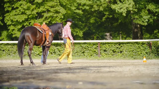 Cowboy lead the horse in riding arena slow mov 4K. Long shot wide view of a male person in focus wearing hat and red shirt while leading a beautiful horse.