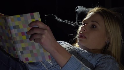 A young beautiful woman lies on a couch in a dark room and reads a book, immersed - closeup