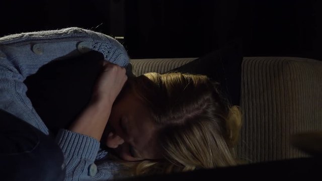A young beautiful woman lies on a couch and cries into a pillow in a dark room - closeup