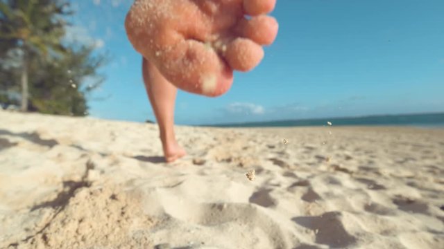 SLOW MOTION, LOW ANGLE, CLOSE UP, DOF: Active girl on holiday jogs barefoot along the sunny tropical coast. Unrecognizable young woman running in the soft sand on a perfect summer day at the seaside.