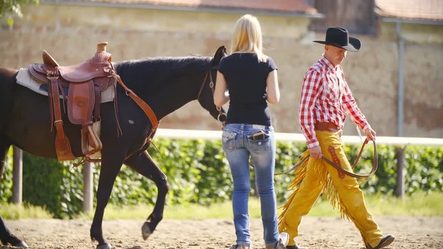 Cowboy and horse walk around a blonde woman in slow motion 4K. Long shot tracking cowboy in focus leading horse passing the blonde woman inside the riding arena.