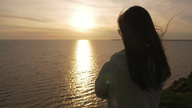 Back view of an attractive girl with long loose hair in a white hoodie standing on the Black Sea shore and looking at golden sunset path in summer