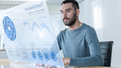 In the Near Future Successful Trader Works on Computer with Transparent Display that Shows Interactive Line Charts and Other Useful Information. His Office is Bright and Modern.