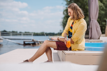 smiling young man in sunglasses holding cocktail and using laptop at poolside
