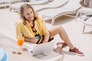 high angle view of smiling young man using laptop while resting with cocktail at poolside
