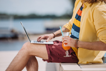 cropped shot of young man holding cocktail and using laptop outdoors