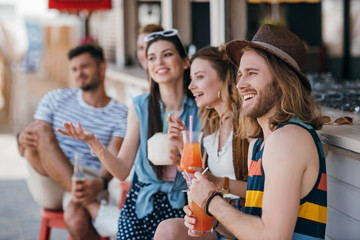 smiling young friends drinking cocktails and looking away at beach bar