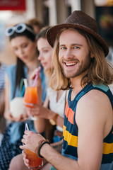 handsome young man smiling at camera while drinking cocktail with friends at beach bar