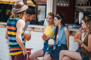 happy young men and women talking and drinking cocktails at beach bar