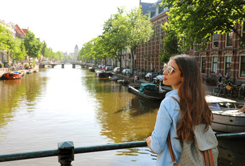 Portrait of traveler girl with sunglasses and backpack enjoying Amsterdam city. Back view of young woman looking to the side on Amsterdam channel, Netherlands, Europe.