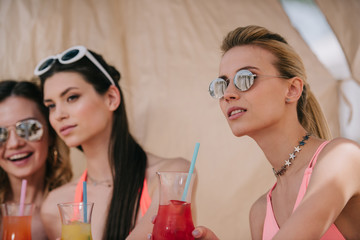 beautiful girls in swimwear and sunglasses drinking cocktails in bungalow on beach