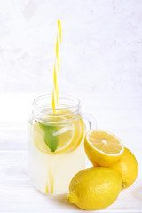 One mason jar glass of homemade refreshing lemonade with slices of organic ripe lemon, whole and halved. Non alcoholic beverage on a rustic white wooden background. Close up, top view, copy space.