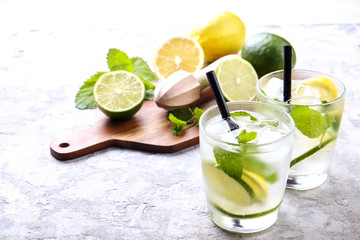 Two glasses of refreshing non alcoholic mojito lemonade drink with organic lemon, lime slices, mint leaves, straw, ice cubes on grunged grey concrete table background. Close up, top view, copy space.