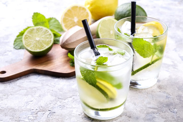 Two glasses of refreshing non alcoholic mojito lemonade drink with organic lemon, lime slices, mint leaves, straw, ice cubes on grunged grey concrete table background. Close up, top view, copy space.