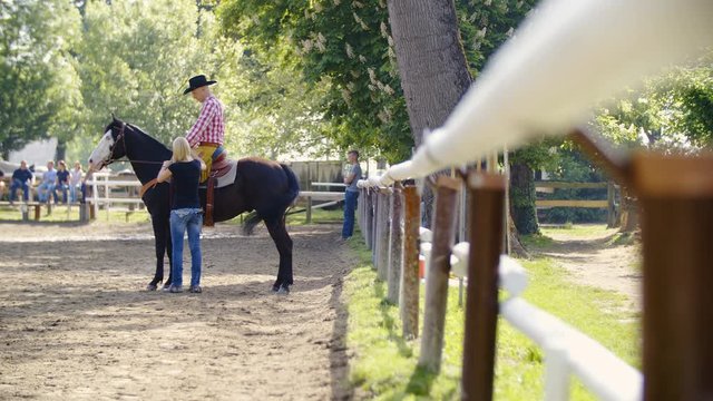 Woman give instructions to cowboy on horse in riding arena 4K. Wide view slow motion of cowboy on a black horse in focus with a blonde girl standing beside. Giving instructions for riding.