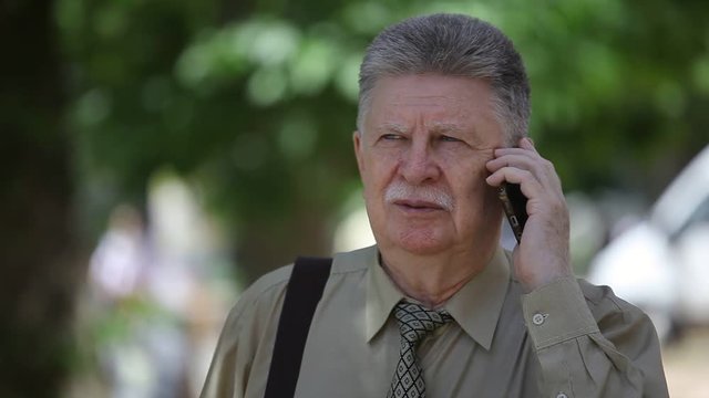 Portrait of an uneasy old man with mustashe talking on phone with his relatives and looking concerned in a picturesque alley on a sunny day in summer