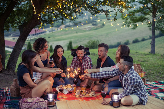 Group of happy millennials have fun with fire sparkles at picnic in spring at sunset on a camping holiday. They drinking wine and eating meat prepared at the barbecue