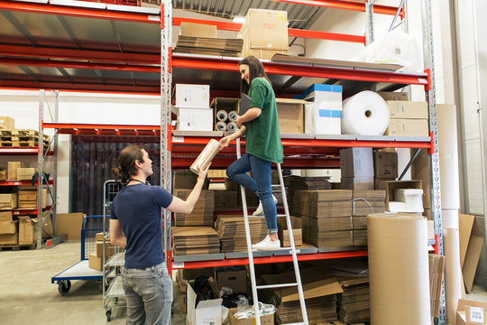 Full length of woman giving rolled up cardboard to male coworker in industry