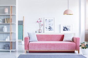 Lamp above pink settee with pillows in pastel living room interior with flowers and posters. Real photo