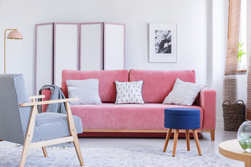 Blue stool next to pink couch in bright living room interior with armchair and poster. Real photo