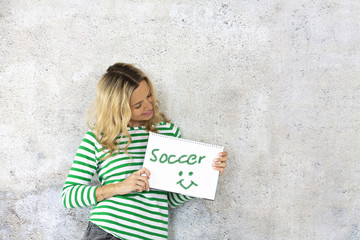 young pretty woman with striped pullover holding checkered paper block in the camera with the word soccer written on it