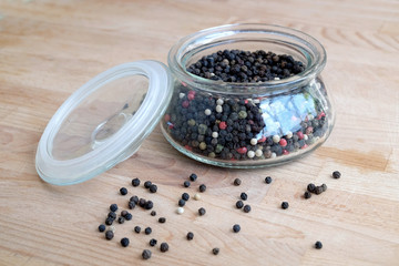 Hot spice ingredients for food. Still life with black pepper seeds inside round glass jar and scattered on wooden table background sde view closeup