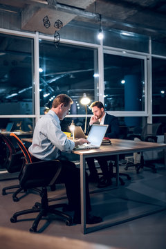 Couple Of Colleagues Sitting At Office Table With Laptops. Two Serious Businessmen Working In Middle Of Night In Their Modern Office With Glass Windows.