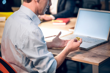 Senior man sitting in front of computer with bitcoin. Cropped picture of guy wearing blue shirt holding golden coin in his hands while sitting at wooden table with laptop on it.