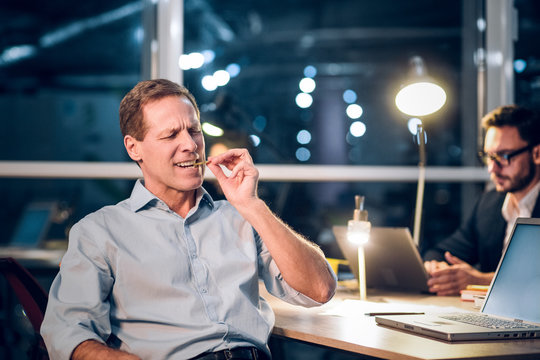 Aged businessman biting bitcoin. Senior man sitting in his office and biting on golden bitcoin with his eyes closed. His colleagues sitting behind him in background working.