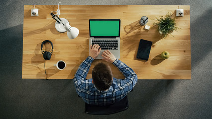 Top View of a Young Creative Man Working on His Green Screen Mock-up Laptop while Sitting at His Wooden Desk. Also on the Table: Coffee Cup, Smartphone, Notebook, Lamp, Plant.