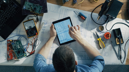 Top View of Competent IT Technician Working on Blueprints, He's Holding Tablet Correcting His Drafts. He's Surround by Circuit Boards, Laptop and Other Technical Components. Sun Shines on His Table.