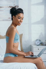 smiling african american girl in lingerie using laptop on bed