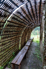A curved bamboo structure with a wooden bench running along the inside about 18" above the bare ground. The weaving is open and you can see the light coming in. The image is take from the open end.
