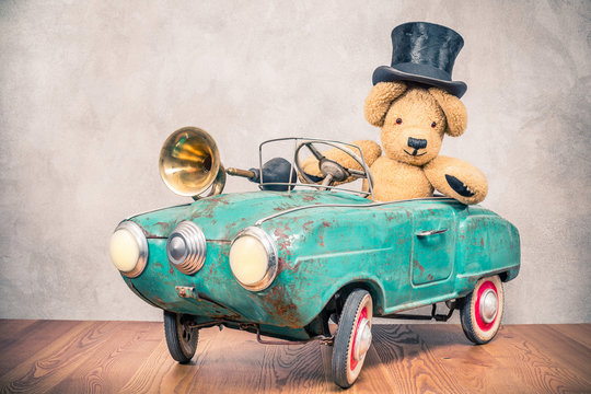 Teddy Bear in old antique cylinder hat driving rusty retro turquoise toy pedal car from circa 60s with classic brass klaxon in front concrete textured wall background. Vintage style filtered photo