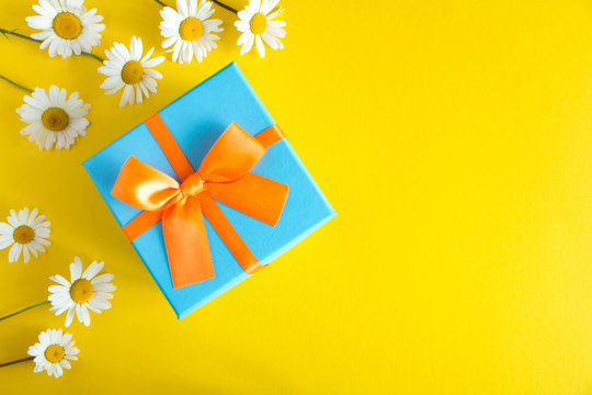 Blue gift box with orange  ribbon and chamomile  on the  yellow background.Top view.