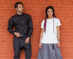 Youth street fashion. Black overalls on a young fashionable man and a beautiful girl in a white T-shirt and a gray skirt with braids posing near a brick wall.