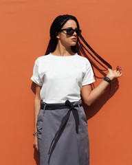 Fashion of youth. Street style. A beautiful girl with African pigtails posing in a fashionable outfit, t-shirt and skirt on an orange wall in the street.