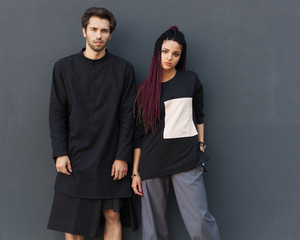 Young beautiful couple girl and young man in trendy not ordinary youth clothes from designer pose against gray wall in city