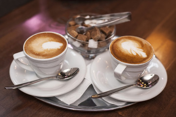 Two white cups of cappuccino are on the tray at the cafe. Behind them stands a bowl of white and brown sugar