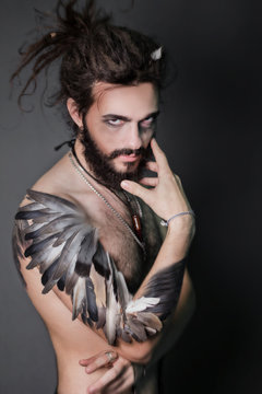 The image for halloween. A brutal guy with dreadlocks on a gray background. The guy with a thick beard. Hand the bird's wing. Feathers on hand. Fantasy