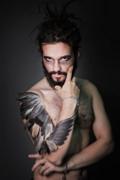 The image for halloween. A brutal guy with dreadlocks on a gray background. The guy with a thick beard. Hand the bird's wing. Feathers on hand. Fantasy