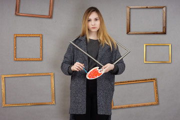 Young artist with palette and picture frames in grey coat on grey background